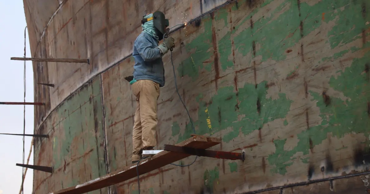 Welder in a helmet standing on a plank and welding the side of a large steel plate structure, a heavily restrained joint of the kind that calls for Z35 steel plate